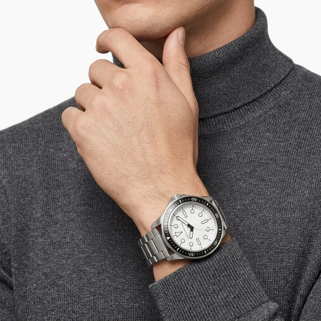 A silver stainless steel wristwatch with a white dial and black bezel, worn on a man's wrist. The man is wearing a dark gray ribbed turtleneck sweater, and the background is a clean white studio backdrop.