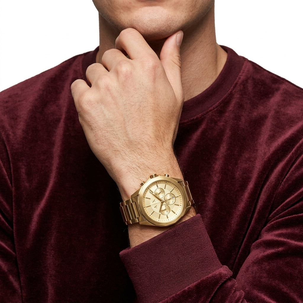 A gold-tone men's watch with a matching metal link bracelet, shown on a model's wrist. The model is wearing a deep burgundy velvet long-sleeve shirt against a clean white background.