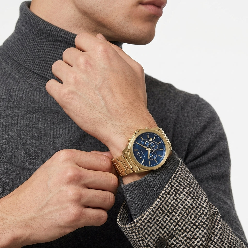 A close-up of a man wearing a gold-tone watch with a blue dial and matching blue bezel, paired with a dark gray turtleneck sweater against a light gray background.
