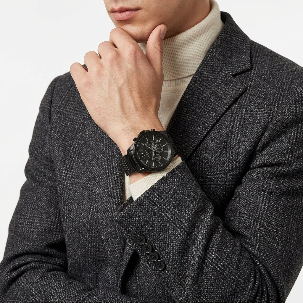 A close-up of a man wearing a black chronograph watch with a black strap on his wrist, styled with a dark gray textured blazer and a cream turtleneck sweater.
