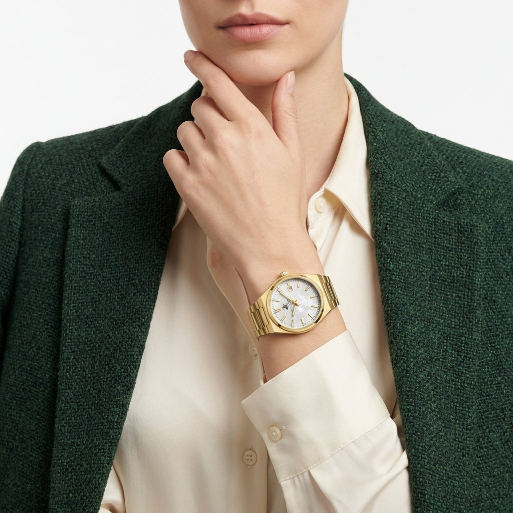 A gold-tone watch with a white dial and bezel, worn on a model's wrist. The model is wearing a cream-colored collared shirt and a dark green textured blazer against a clean white background.