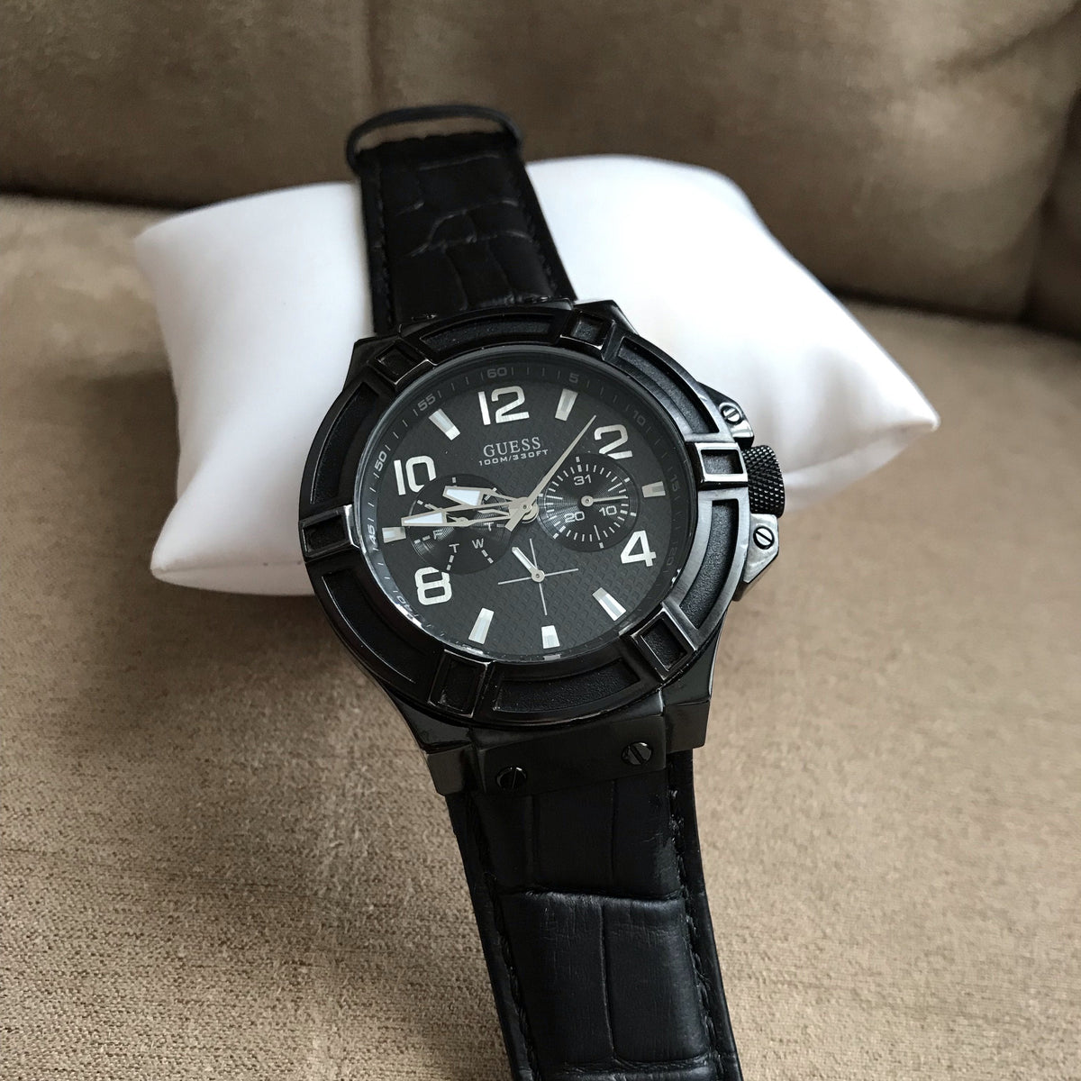 Guess Rigor watch displayed on white pillow showing black dial and leather strap with croc-embossed texture