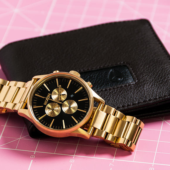 Nixon Sentry Chrono gold watch with black wallet on pink textured background