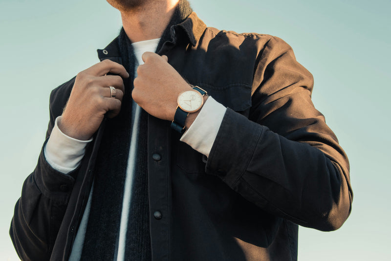 Person wearing a black jacket with a white shirt and a watch, against a light blue background