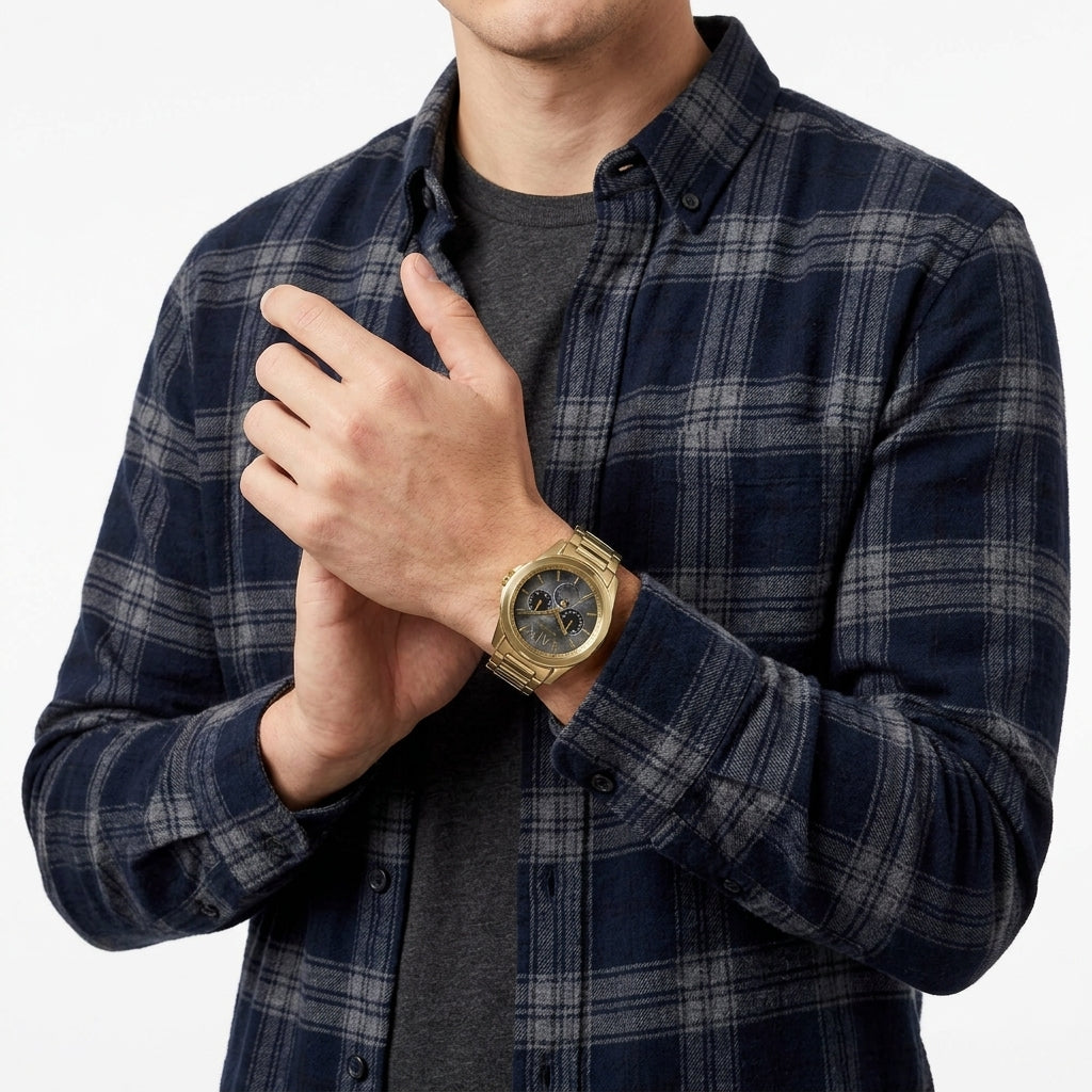 A man wearing a navy and gray plaid button-down shirt over a dark gray t-shirt, with a gold-tone wristwatch on his left wrist, against a plain white background.