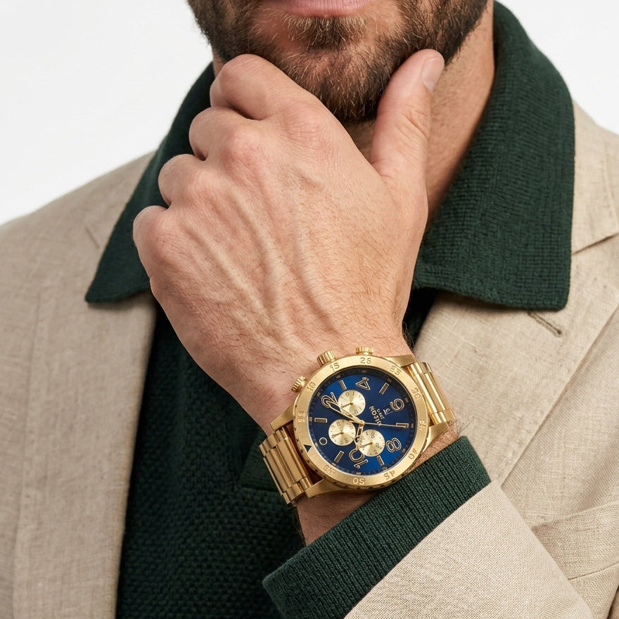 A close-up lifestyle shot of a man wearing a gold-tone wristwatch with a blue dial and gold bracelet, styled with a dark green knit collar shirt and a beige blazer.