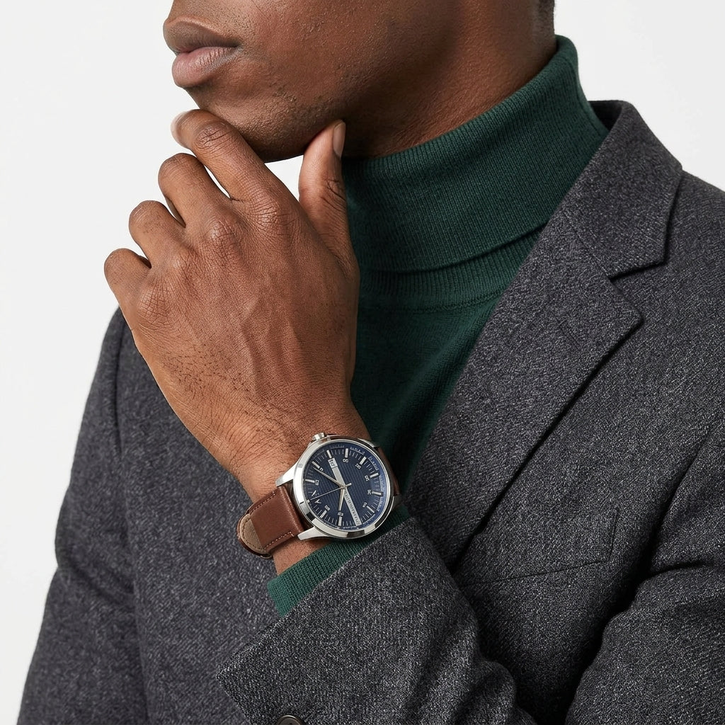 A close-up of a man wearing a brown leather strap watch with a dark blue dial and silver-tone case, styled with a dark green turtleneck and a textured gray blazer.