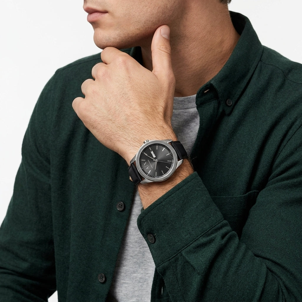 A close-up of a man wearing a silver-toned smartwatch with a black dial and black strap, dressed in a dark green button-up shirt over a gray t-shirt, against a clean white background.