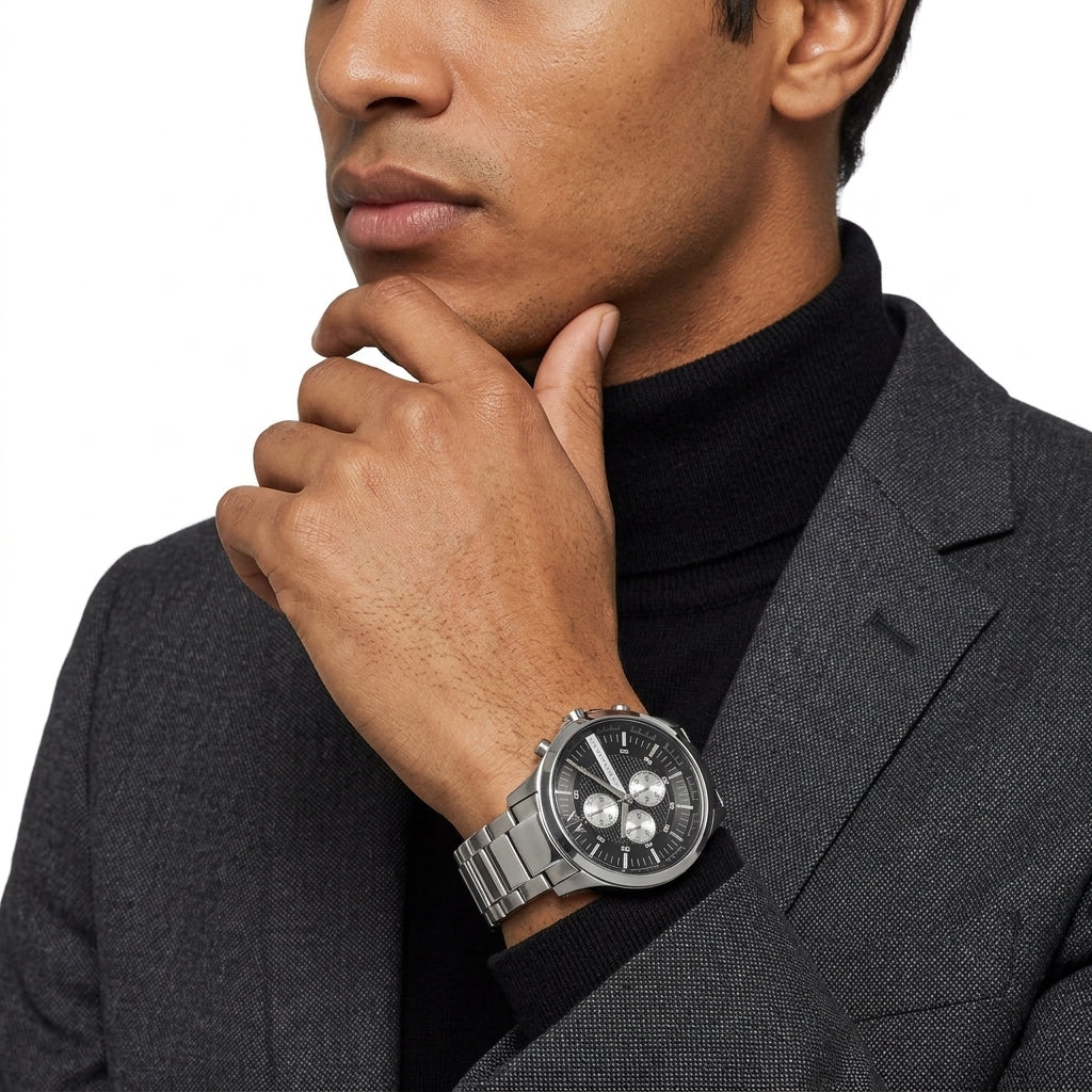 A close-up of a man wearing a silver stainless steel watch with a black dial and silver link bracelet, styled with a black turtleneck and gray blazer against a white background.