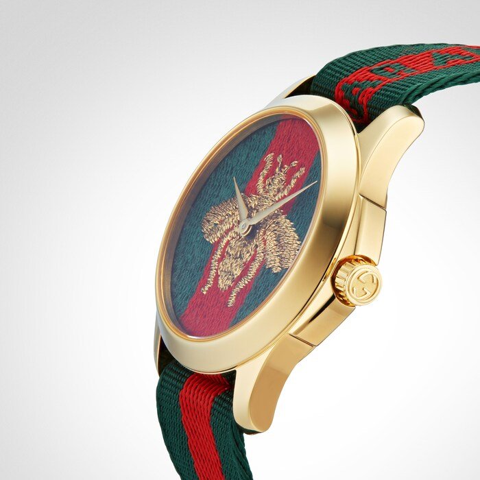 Side angle view of Gucci G-Timeless watch showing gold case, bee motif dial, and striped fabric strap